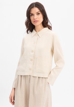 Vrouw met golvend bruin haar draagt een crèmekleurige jas met knopen en twee voorzakken en beige broek met hoge taille tegen een effen witte achtergrond.