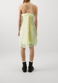 Světle žluté vrstvené šaty s texturovaným povrchem, nastavitelné tenké ramínka a detailem na zádech. Kombinováno s černými kotníkovými botami.