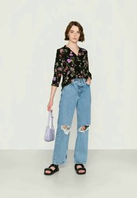 Camisa preta com padrão floral, jeans rasgados azuis claros, sandálias pretas e uma pequena bolsa púrpura, tudo em um fundo neutro.