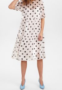 Genomskinlig klänning med prickar i ljus rosa med svarta prickar. Har en lagerdesign, korta ärmar och en avslappnad passform. Bärs med ljusblå klackar.