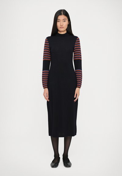 Langes schwarzes Kleid mit tailliertem Oberteil und mittellanger Länge. Hat gestreifte Ärmel in Rot und Orange. Aus glattem Strickstoff, mit Rundhalsausschnitt, getragen mit schwarzen Ballerinas.