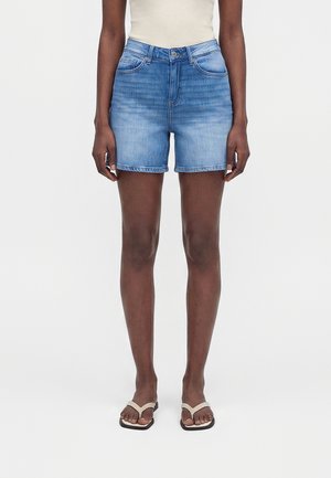 Kvinde iført blå denimshorts med mellemhøj talje, hvid ærmeløs top og beige sandaler, stående mod en ensfarvet hvid baggrund.
