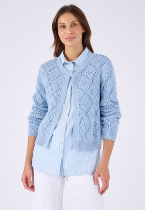 Donna che indossa un cardigan lavorato a maglia azzurro chiaro con motivi a diamante sopra una camicia a righe blu e bianche con bottoni e pantaloni bianchi.