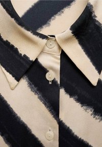 Col de chemise crème avec boutons blancs et larges rayures diagonales noires inégales sur le tissu.