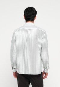 Camisa de manga larga en rayas verticales de color gris claro y blanco, que presenta un cuello con botones y una abertura trasera con un detalle de bucle.