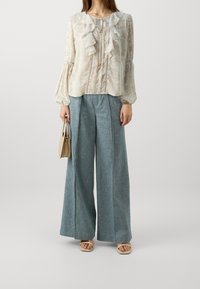 Femme portant un chemisier ivoire à volants, un pantalon large bleu, des talons ouverts beige et tenant un sac à main beige devant un fond uni.