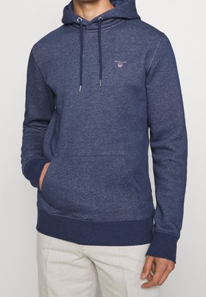 Mann trägt einen marineblauen GANT-Hoodie mit Fronttasche und Kordeln, kombiniert mit hellen Hosen.