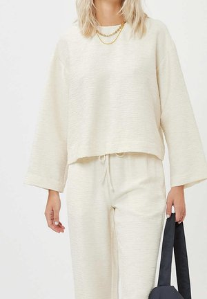 Femme portant un haut ample crème à manches longues et un pantalon, tenant un sac cabas noir, avec des colliers en or superposés visibles.