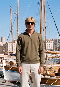 Homme blond portant des lunettes de soleil, un pull vert olive et un pantalon blanc, debout devant des voiliers en bois dans une marina ensoleillée.