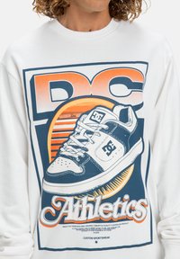 Witte sweatshirt met een grafisch ontwerp featuring een blauwe en oranje illustratie van een sneaker en de tekst "DC Athletics."