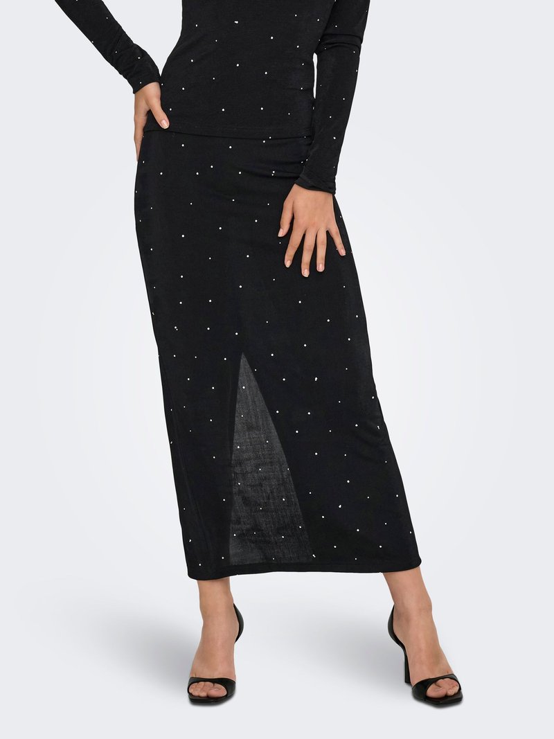 Femme portant une jupe midi noire avec de petits pois blancs et une fente avant, associée à des talons ouverts noirs sur fond blanc.