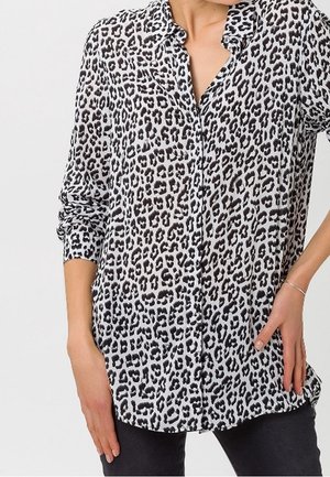 Kobieta ubrana w koszulę na guziki z długim rękawem w czarno-biały wzór w panterkę, do tego czarne spodnie, ręka oparta na biodrze.