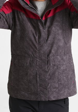 Osoba ubrana w ciemnoszary płaszcz z czerwonymi akcentami, dużymi przednimi kieszeniami i zamkiem błyskawicznym przykrytym patką.