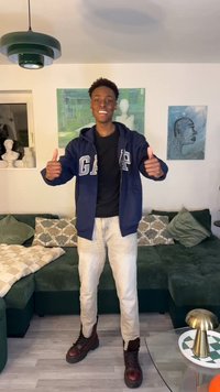 Ung mann smiler og gir tommel opp, stående i stuen med grønn sofa, dekorative puter og moderne kunstverk på veggene.