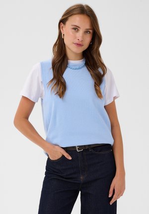 Femme aux longs cheveux bruns portant un pull sans manches bleu clair sur un t-shirt blanc et un jean foncé, une main dans la poche, debout, fond neutre.