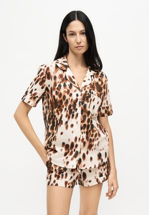 Femme aux longs cheveux foncés porte un haut boutonné à manches courtes et un short assorti avec un motif tacheté marron et noir sur un tissu clair.