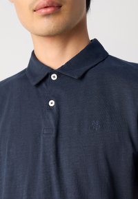 Tamsiai mėlynos polo marškiniai pagaminti iš tekstūrinio audinio su klasikinėmis apykaklėmis. Yra dvi baltos mygtukai ir siuvinėtas logotipas kairėje krūtinės pusėje.