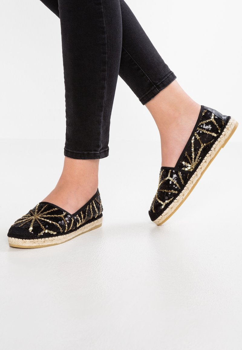 Czarne espadryle z złotymi cekinami, okrągły nos i pleciona juta na podeszwie. Noszone z dopasowanymi czarnymi dżinsami, ukazujące profil boczny.