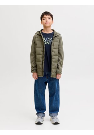 Junge steht und trägt eine olivgrüne Kapuzenjacke, ein marineblaues T-Shirt mit Grafikdruck, lockere blaue Jeans mit umgeschlagenen Bündchen an den Knöcheln und weiße Turnschuhe vor weißem Hintergrund.