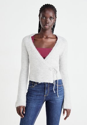 En ung kvinde med fletning bærer en lys grå omslagstrøje over en maroon top og blå jeans, stående mod en hvid baggrund.