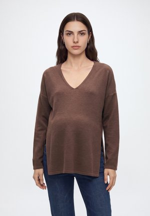 Femme enceinte aux longs cheveux foncés portant un pull marron à col en V et un jean bleu, debout devant un fond blanc uni.