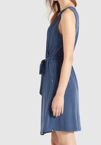 Femme portant une robe bleu marine sans manches avec une ceinture à la taille, debout de profil devant un fond uni gris clair.