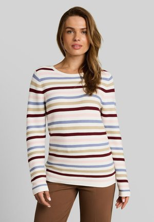 Vrouw met lang bruin haar die een gestreepte trui met lange mouwen draagt in beige, blauw, bordeaux en roze met een bruine broek, staand tegen een grijze achtergrond.