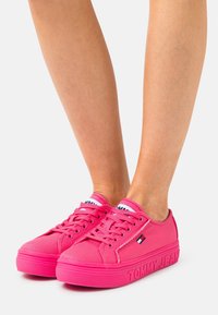 Zapatillas de lona rosas con suelas planas, punteras redondas y diseño con cordones. Presenta un logo en la lengüeta y el costado, y una suela de goma texturizada.