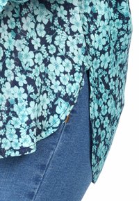 Floral blouse with a navy background, featuring light blue flowers. Fabric is lightweight with a rounded hem and side slits. Worn over blue jeans.
