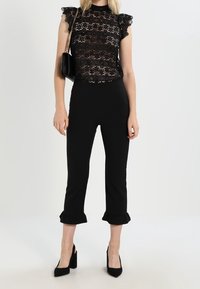 Musta pitsi toppi, jossa on röyhelöhiat, yhdistettynä mustiin slimmeihin housuihin, joissa on röyhelöreunat. Mallilla on pieni musta laukku ja korkokengät.