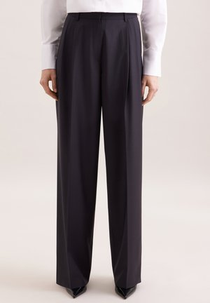 Pantalon large noir, taille ajustée, porté avec une chemise blanche rentrée, associé à des chaussures noires à bout pointu, sur un fond neutre.