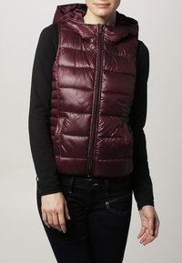 Bordeauxfarbene gesteppte Daunenweste mit Kapuze, vorn mit Reißverschluss, getragen über einem schwarzen Langarmshirt.