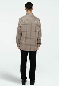 Chemise à carreaux marron en tissu doux, avec des manches longues, un col et des coutures noires. Portée avec un pantalon noir et des bottes.