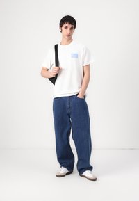 White t-shirt with blue graphic text, paired with loose dark blue jeans and sneakers. Black shoulder bag hangs from one shoulder.