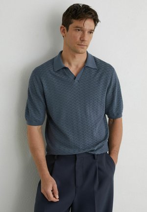 Mężczyzna o ciemnych włosach, ubrany w niebieską, teksturowaną koszulkę polo z krótkim rękawem i granatowe spodnie, stojący oparty o białą ścianę, z rękami w kieszeniach.