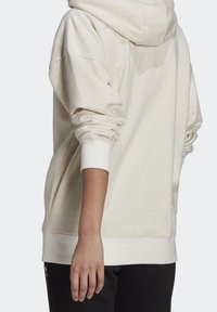 Osoba ve světle béžové mikině s vyhrnutými rukávy a černými kalhotami, zobrazená zezadu a ze strany.