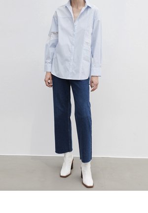 Vrouw draagt een lichtblauw gestreept overhemd met knopen, korte donkerblauwe wijde jeans en witte enkellaarsjes met hak, staand op een grijze vloer.