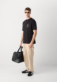 Must must t-särk logotrükiga, beežid laiad püksid, must nahast spordikott ja mustad sneakers valgete triipudega.