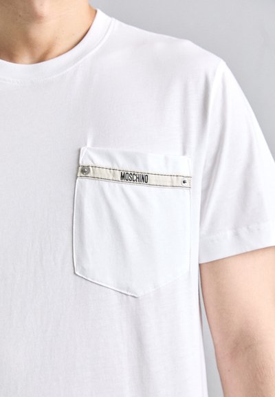 Suurendatud vaade valge t-särgi taskust, millel on beeži ääris, metallist nööbid ja must "MOSCHINO" logo tekst.