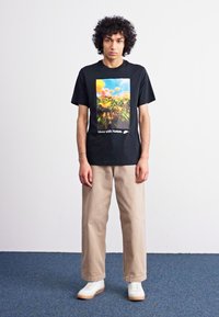 Camiseta gráfica preta com uma cena da natureza com flores laranja, combinada com calças bege e ténis brancos com solas de borracha.