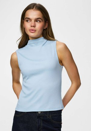 Kobieta w jasnoniebieskiej bluzce bez rękawów z wysokim kołnierzem i ciemnych dżinsach, pozująca z rękami za plecami, patrząca w prawo.