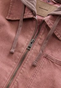 Gros plan sur une veste en denim rose poussiéreux avec une fermeture éclair métallique, des cordons de serrage, un col et des coutures visibles sur la poche poitrine.