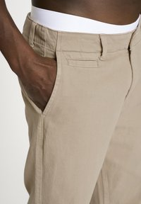 Beige bomuldsbukser med struktureret stof, sidelommer og en elastisk hvid talje. Pasformen er designet til afslappet brug.