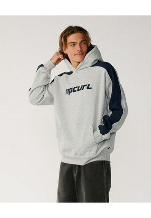 Młody mężczyzna noszący szarą bluzę z kapturem Rip Curl z granatowymi paskami na rękawach, stoi z jedną ręką za głową, a drugą w kieszeni.