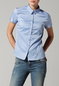 Chemise bleu clair à manches courtes avec col, fermeture à boutons et un subtil détail de fronces à l'avant, associée à un jean en denim déchiré.