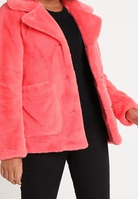 Faux fur, bright pink coat with a notched collar, front pocket, and button closure. Worn over a black knit sweater. Soft texture.