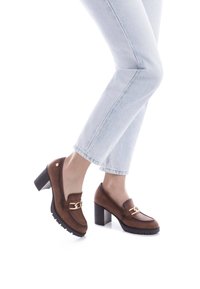 Mocasines marrones de tacón con un acento de cadena, que cuentan con un exterior de cuero suave, punta redondeada y suela de goma texturizada. Llevados con jeans de mezclilla claros.