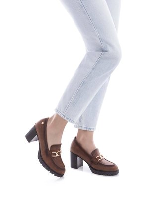 Mocasines marrones de tacón con un acento de cadena, que cuentan con un exterior de cuero suave, punta redondeada y suela de goma texturizada. Llevados con jeans de mezclilla claros.
