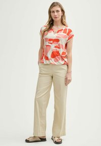 Haut à manches courtes en crème avec des formes abstraites rouges et roses ; associé à un pantalon large beige et des sandales brunes.