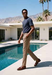Homme portant des lunettes de soleil, un t-shirt bleu marine, un pantalon à motifs et des mocassins marron, marchant à côté d'une piscine avec des montagnes et des palmiers en arrière-plan.